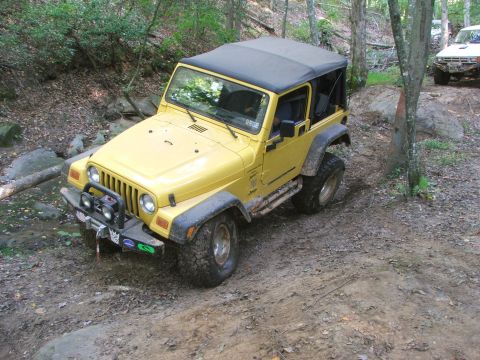 1996 Ford Bronco - A yellow Jeep Wrangler navigates a wooded off-road trail, with another vehicle visible in the background.