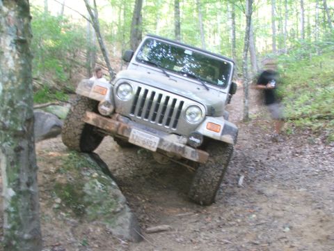 1996 Ford Bronco - A silver Jeep Wrangler navigates a challenging off-road trail with large rocks and trees.