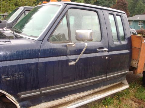 1989 Ford Bronco - A dark blue pickup truck is parked next to another vehicle, with a wooden flatbed visible on the right side.