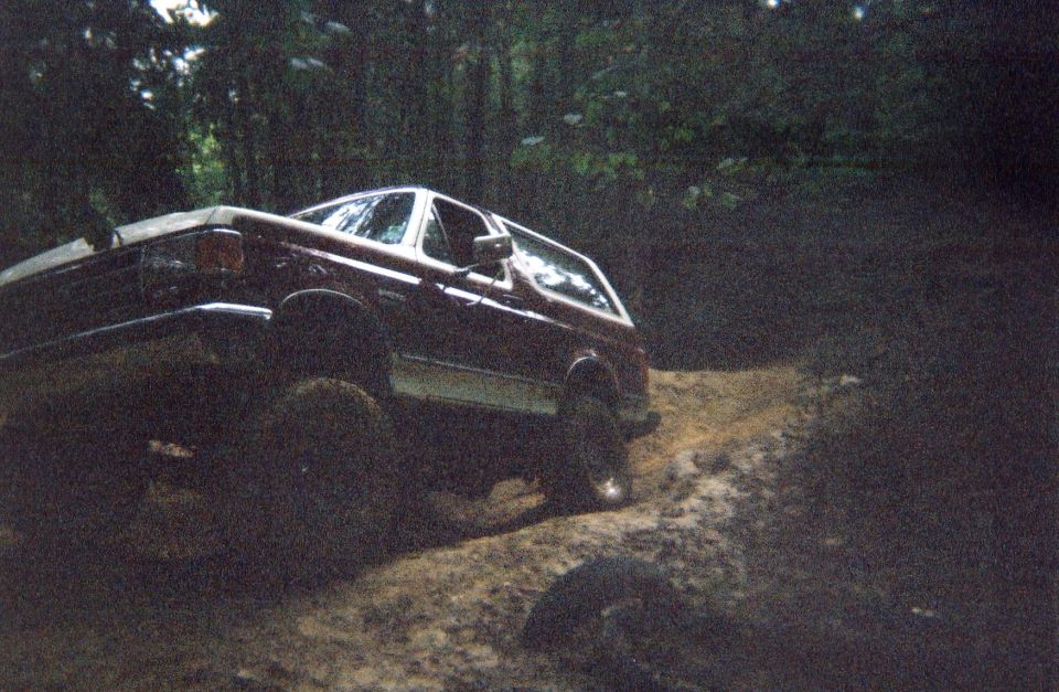 Dark red lifted SUV flexing on an off-camber dirt mound in wooded trail
