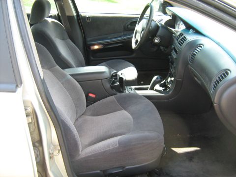 1988 Mercury Grand Marquis - Interior of a silver sedan shot through the open passenger door, showing gray cloth bucket seats, a center console with cupholders and floo…