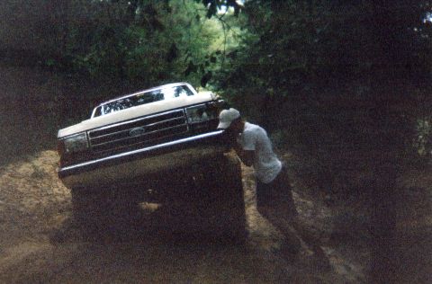 1990 Ford Bronco - A man pushes a white Ford Bronco as it navigates a muddy, off-road trail.