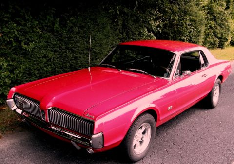 1967 Mercury Cougar - A red 1967 Mercury Cougar is parked on a paved surface with a hedge behind it.