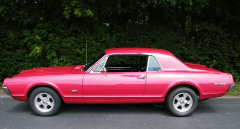 1967 Mercury Cougar - A red 1967 Mercury Cougar coupe is shown from the side against a backdrop of green foliage.