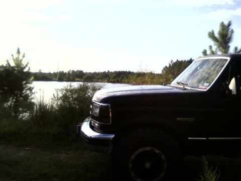 1994 Ford Bronco - A dark brown Ford Bronco is parked near a body of water under a bright sky.