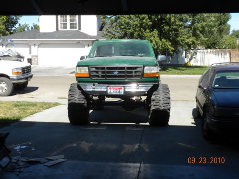 1989 Ford Bronco - A lifted green truck with oversized tires is parked in front of a garage, with another truck and a sedan visible.