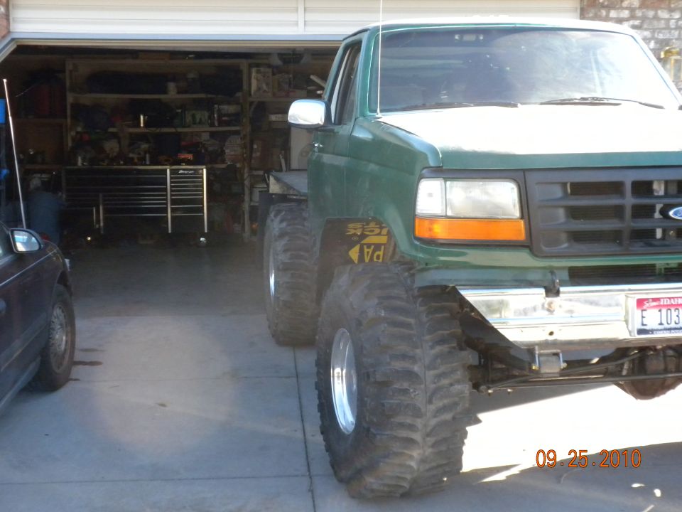 Green pickup truck with big tires parked in a garage next to a dark car