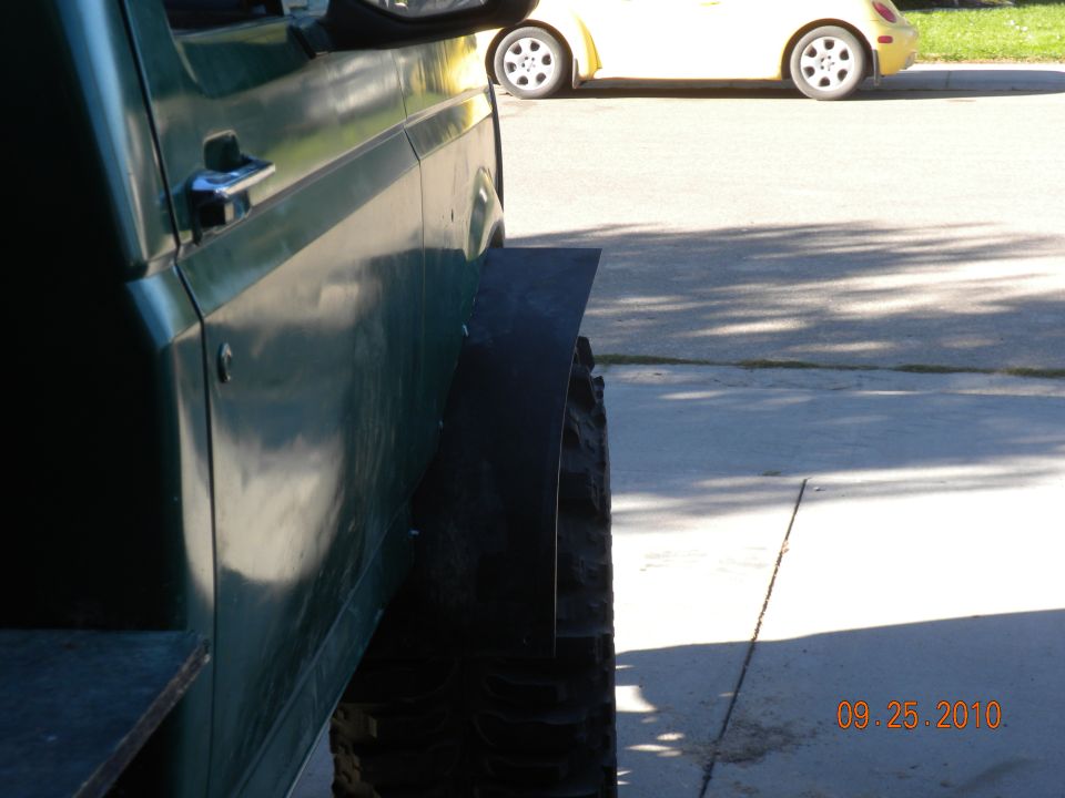 Side view of a green SUV with oversized tires and fender flares. A yellow car is parked in the background