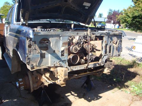 1989 Ford Bronco - Blue Ford truck stripped down to the bare radiator support and inner fenders, sitting on jack stands in a driveway with the front wheels of…