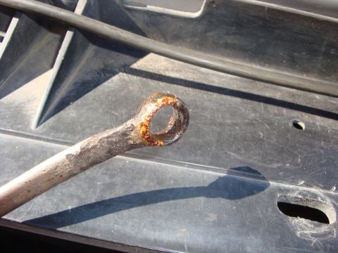 1989 Ford Bronco - Sunlit close-up of a heavily rust-pitted closed ring end on a metal rod, its bore crusted with orange corrosion, resting on a scratched bla…