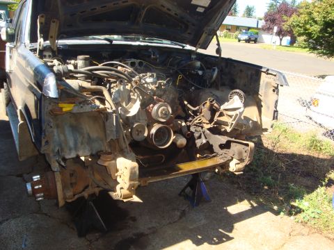 1989 Ford Bronco - Mid-teardown blue Ford truck sitting on blue jack stands in a driveway, front sheet metal, radiator and one fender removed to expose the V8…