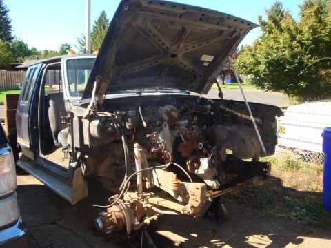 1989 Ford Bronco - A dark blue Ford Bronco stripped down mid-build in a sunny driveway, front fenders and grille removed with the hood propped open over a bar…