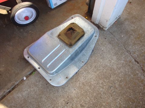 1989 Ford Bronco - A bare stamped-steel transmission pan sits upside down on a concrete driveway, its zinc-plated surface showing three raised ribs, a bolt-ho…