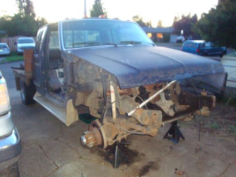 1989 Ford Bronco - Blue extended-cab flatbed Ford truck torn down on jack stands in a driveway with both front fenders, inner fenders and grille removed, expo…
