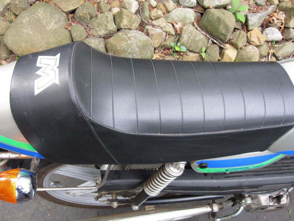 Black ribbed motorcycle seat with white M logo on a silver frame with blue and green stripes