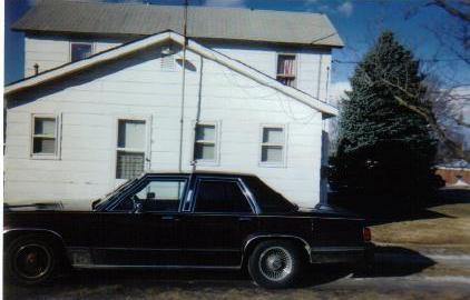 1990 Mercury Grand Marquis - Side view of a dark-colored 1980s Mercury sedan parked outside a house.
