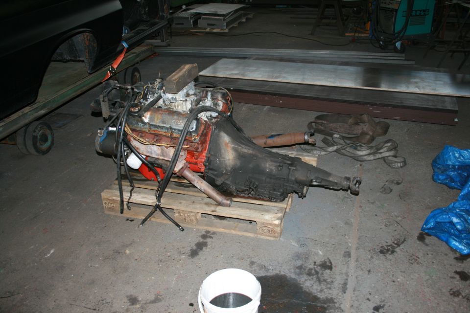 Engine and transmission on a wooden pallet, with part of a black car body on a trailer in the background