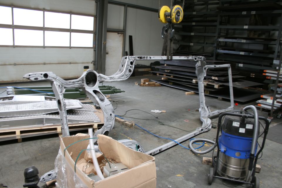 Stripped bare metal vehicle frame on blocks in a workshop with chain hoist, sheet metal racks, and shop vac.