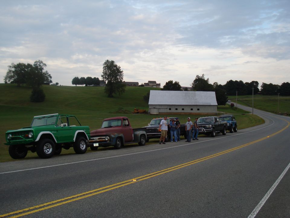 Green Ford Bronco, red Ford F-100, black Ford F-150, and blue Ford Bronco parked on roadside with people