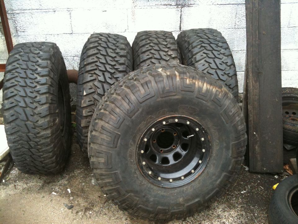 Five mud-terrain tires by a block wall, front one on a black beadlock-style steel wheel