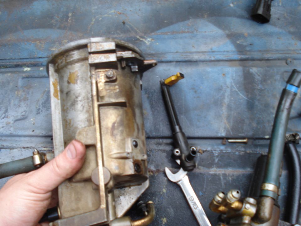 Hand holding dirty metal fuel filter housing with attached hoses and a wrench nearby
