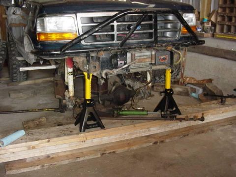 1992 Ford Bronco - Front view of a dark green 1990s Ford truck up on yellow jack stands in a garage with its front axle pulled out, exposing the bare frame ho…
