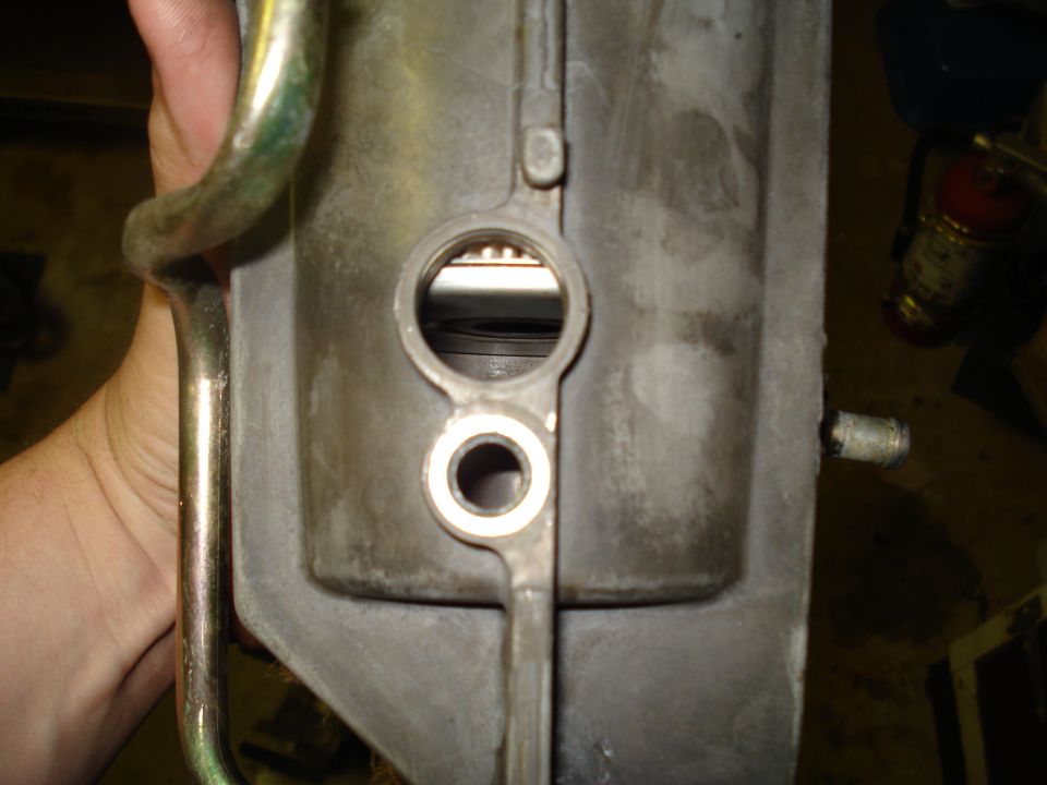 Gray metal engine component with two circular openings, one above the other. A hand holds a metal pipe