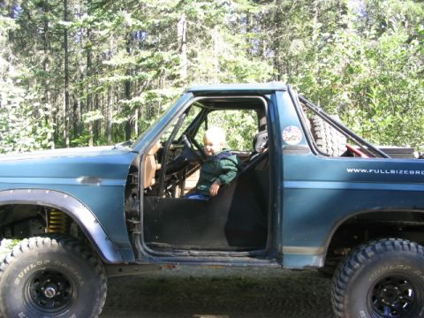 1986 Ford Bronco - A young child sits in the driver's seat of a modified blue SUV, surrounded by a forest.