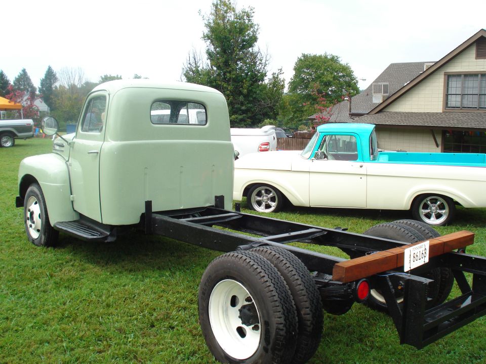 Light green truck chassis with cab, rear view. White truck with blue bed in background