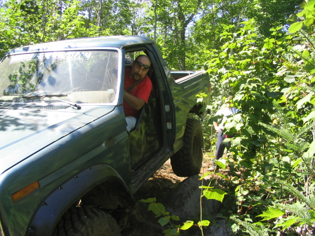 Green SUV driving over rocks on a wooded trail. Driver in red shirt visible