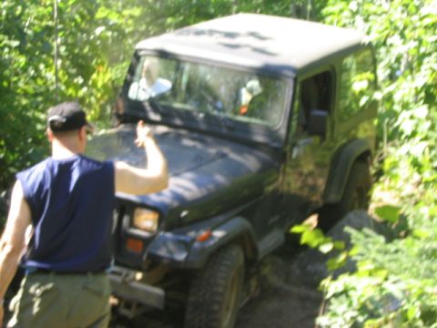 1986 Ford Bronco - A dark Jeep Wrangler navigates a wooded trail with a driver giving a thumbs up from inside.