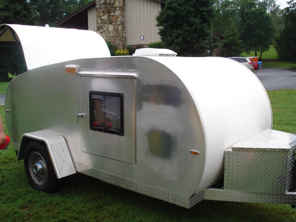 Teardrop trailer with a white curved roof and a silver body. It has one door with a small window