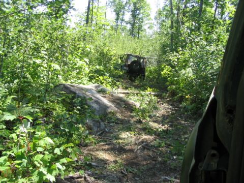 1986 Ford Bronco - Two vehicles navigate a wooded trail with dense foliage and a large rock.