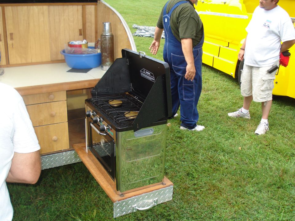Outdoor kitchen setup with a propane stove and oven, two men standing nearby, and a yellow vehicle in the background