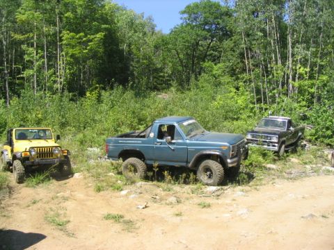 1986 Ford Bronco - Three off-road vehicles, including a blue pickup and a yellow jeep, are parked on a dirt trail surrounded by trees.