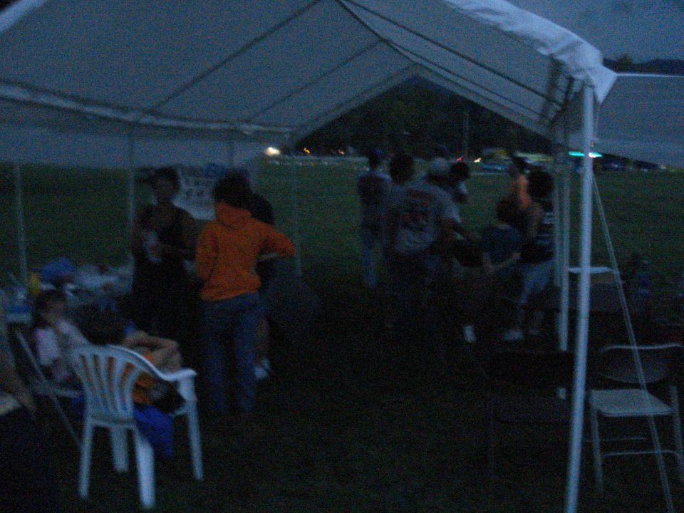 People gathered under a white tent at dusk, with a grassy field in the background