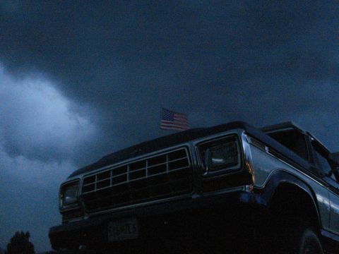 1994 Ford F-350 - A dark 1979 Ford F-Series truck is seen from the front, with an American flag mounted on its roof, under a dramatic, cloudy sky.