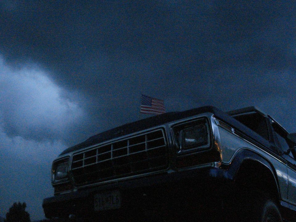 Front view of a dark 1979 Ford F-Series truck with an American flag on top, under a stormy sky