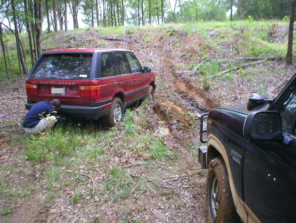 Dark SUV in foreground on muddy trail, red Range Rover stuck behind with person crouching by wheel