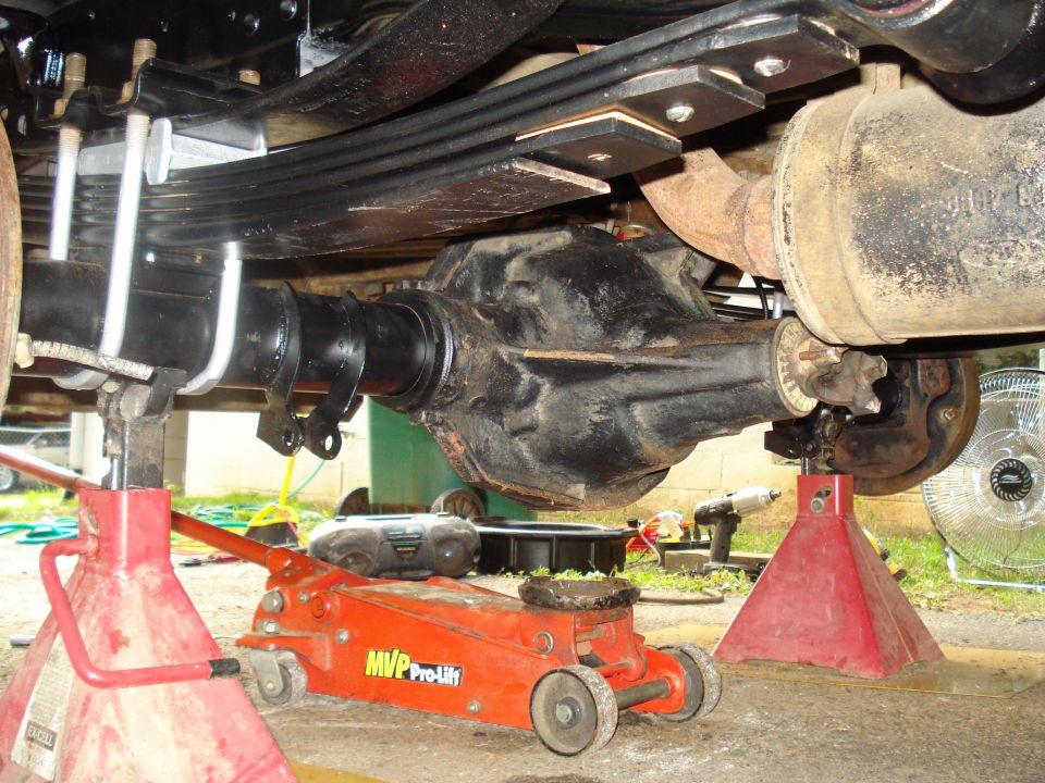 Underside of a vehicle showing the rear axle, leaf springs, and exhaust pipe supported by jack stands and a floor jack
