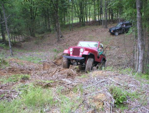 1983 Ford Bronco - Two off-road vehicles navigate a wooded trail with loose dirt and debris.