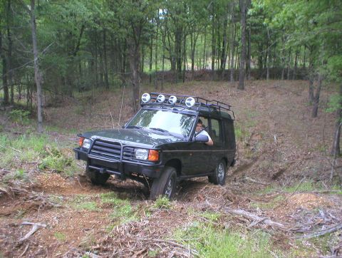 1983 Ford Bronco - A driver steers a dark green Land Rover Discovery I through a wooded area with a roof rack and auxiliary lights.