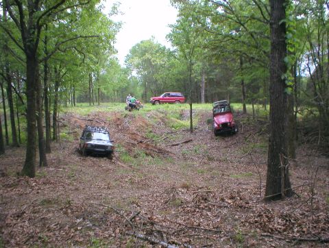 1983 Ford Bronco - Off-road vehicles tackle a challenging dirt track surrounded by trees. Multiple vehicles are visible in the scene.