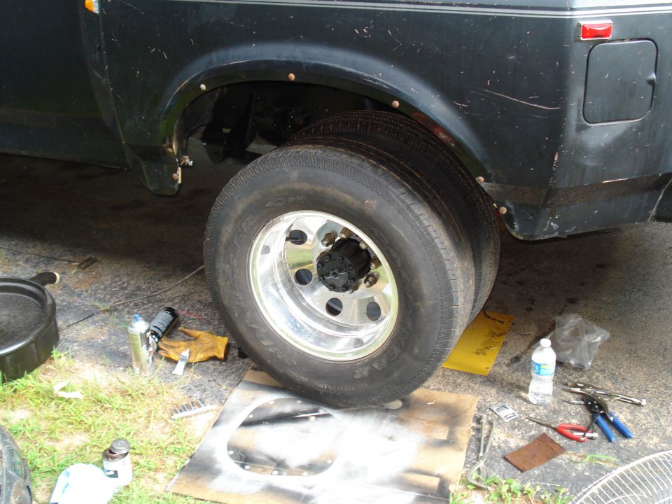 Dual rear wheels on a dark truck, showing chrome rims and tires. Tools and oil pan are on the ground