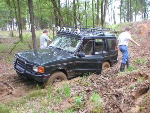 1983 Ford Bronco - A Land Rover Discovery is stuck in a muddy wooded area with two men observing.