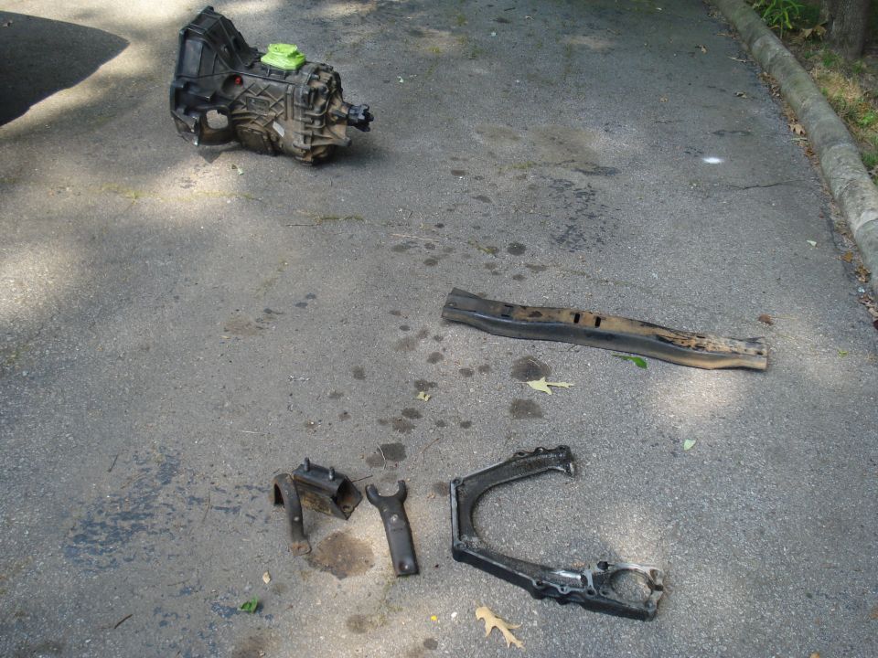 Transmission, control arm, and bracket parts laid out on asphalt