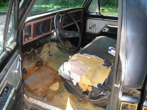 1984 Ford F-250 - The interior of a 1977 Ford F-100 reveals a worn driver's seat, cracked dashboard, and rusty floorboards.