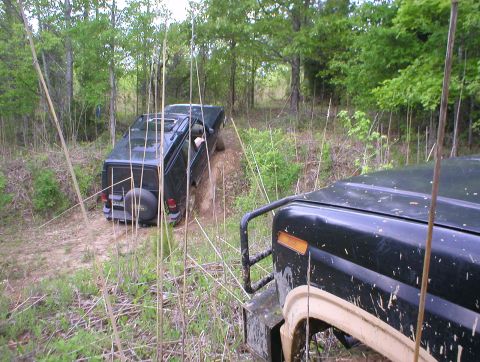 1983 Ford Bronco - Two dark SUVs navigate a wooded trail, one climbing a steep incline.