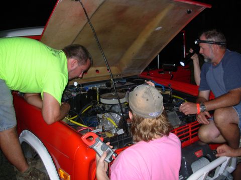 1983 Ford Bronco - A group of people are gathered around the open engine bay of a red vehicle, appearing to work on it at night.