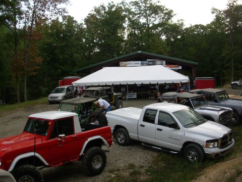 1983 Ford Bronco - A red 1983 Ford Bronco with a white top and off-road tires is parked in a lot with other vehicles and a large tent.
